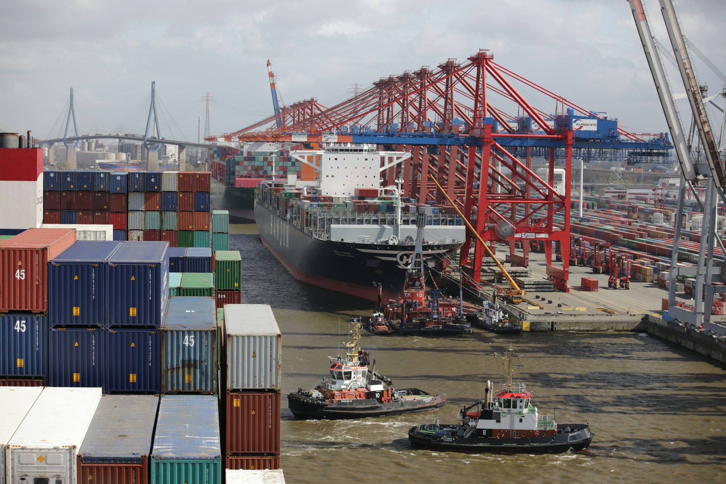 Tugs alongside a container ship under red harbour cranes